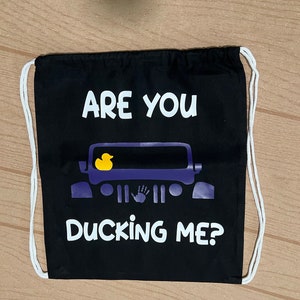 May include: Black drawstring bag with white text that reads "ARE YOU DUCKING ME?" A purple vehicle graphic with a yellow rubber ducky inside is centered on the bag. White drawstring cords are visible.