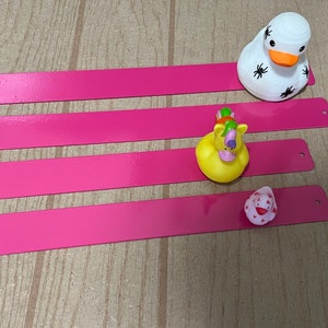 Puede incluir: Cuatro tiras rectangulares rosas con bordes redondeados y agujeros en el lado derecho. Un pato de goma blanco con ara&ntilde;as negras sobre &eacute;l est&aacute; en la tira superior. Un pato de goma amarillo con cabeza de unicornio y un pato de goma rosa con un dise&ntilde;o de coraz&oacute;n rojo est&aacute;n en las tiras inferiores.