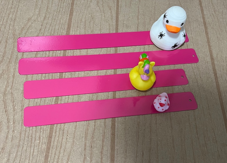 Puede incluir: Cuatro tiras rectangulares rosas con bordes redondeados y agujeros en el lado derecho. Un pato de goma blanco con ara&ntilde;as negras sobre &eacute;l est&aacute; en la tira superior. Un pato de goma amarillo con cabeza de unicornio y un pato de goma rosa con un dise&ntilde;o de coraz&oacute;n rojo est&aacute;n en las tiras inferiores.