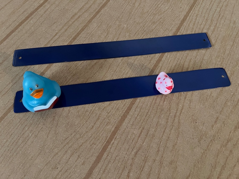 Puede incluir: Dos tiras magn&eacute;ticas azules con un pato azul y un pato rosa con lunares rojos en la parte superior.