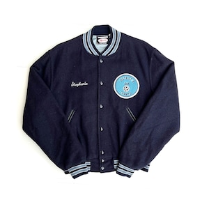 Könnte beinhalten: Eine marineblaue Varsity-Jacke mit einem weißen und hellblauen gestreiften Kragen und Bündchen. Die Jacke hat einen Knopfverschluss und zwei Taschen. Die Jacke hat den Text "Stephanie" auf der linken Brust gestickt und einen Aufnäher auf der rechten Brust, auf dem "District 6 AAA Champs" mit einem Fußball-Motiv steht.