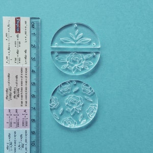 K&ouml;nnte beinhalten: Zwei transparente Acrylkreise mit Blumendesigns. Der obere Kreis ist in zwei H&auml;lften geteilt und zeigt eine gro&szlig;e Blume mit Bl&auml;ttern. Der untere Kreis zeigt ein sich wiederholendes Muster aus kleineren Blumen und Bl&auml;ttern.