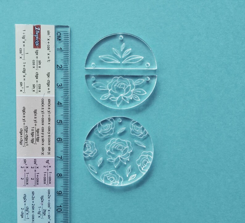 K&ouml;nnte beinhalten: Zwei transparente Acrylkreise mit Blumendesigns. Der obere Kreis ist in zwei H&auml;lften geteilt und zeigt eine gro&szlig;e Blume mit Bl&auml;ttern. Der untere Kreis zeigt ein sich wiederholendes Muster aus kleineren Blumen und Bl&auml;ttern.