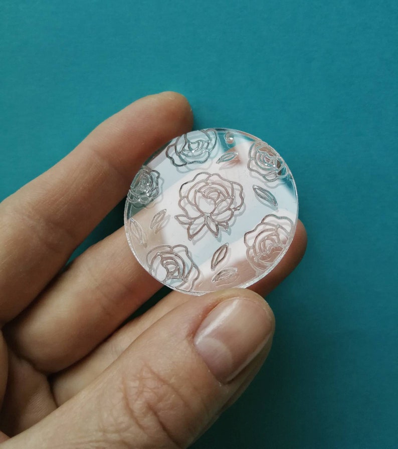 K&ouml;nnte beinhalten: Ein runder, transparenter Acryl-Anh&auml;nger mit einem wei&szlig;en, ge&auml;tzten Blumenmuster mit Rosen und Bl&auml;ttern.
