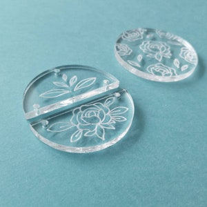 K&ouml;nnte beinhalten: Zwei transparente Acrylkreise mit einem eingegravierten Blumendesign. Die Kreise sind getrennt und durch eine kleine Metallstange verbunden.