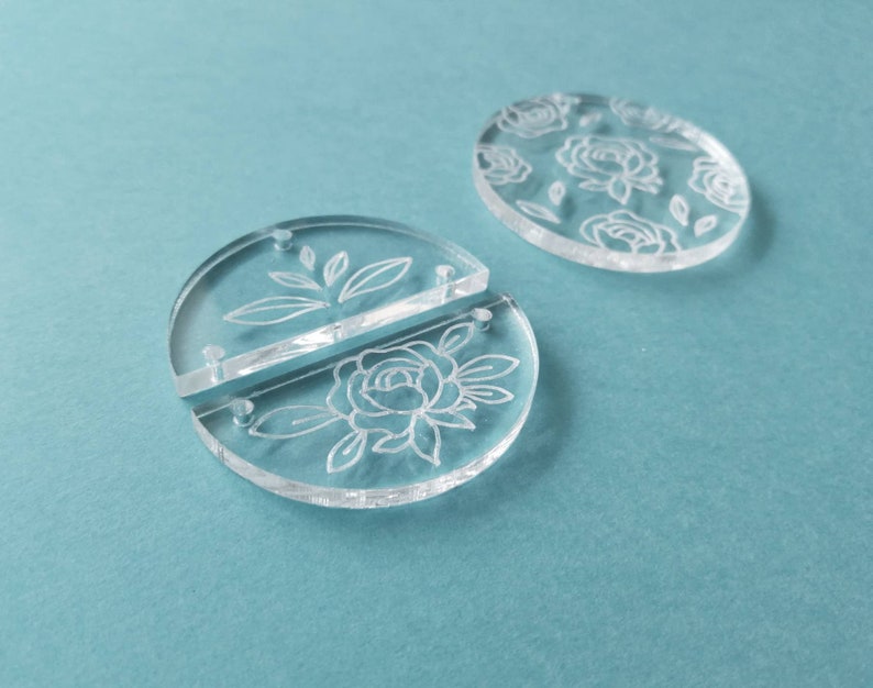 K&ouml;nnte beinhalten: Zwei transparente Acrylkreise mit einem eingegravierten Blumendesign. Die Kreise sind getrennt und durch eine kleine Metallstange verbunden.