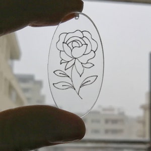 K&ouml;nnte beinhalten: Ein transparenter, ovaler Acryl-Anh&auml;nger mit einer schwarzen Linienzeichnung einer einzelnen Rose mit Bl&auml;ttern.