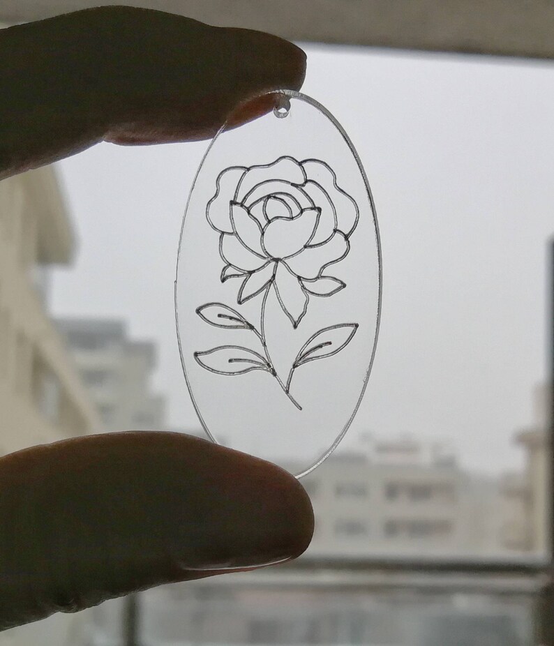 K&ouml;nnte beinhalten: Ein transparenter, ovaler Acryl-Anh&auml;nger mit einer schwarzen Linienzeichnung einer einzelnen Rose mit Bl&auml;ttern.