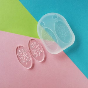 K&ouml;nnte beinhalten: Zwei transparente, ovale Ohrringrohlinge aus Acryl mit einem wei&szlig;en Blumenmuster. Die Rohlinge werden neben einer transparenten Silikonform mit zwei ovalen Vertiefungen gezeigt.