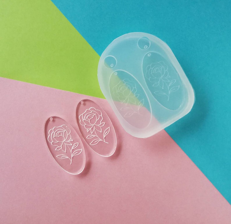 K&ouml;nnte beinhalten: Zwei transparente, ovale Ohrringrohlinge aus Acryl mit einem wei&szlig;en Blumenmuster. Die Rohlinge werden neben einer transparenten Silikonform mit zwei ovalen Vertiefungen gezeigt.