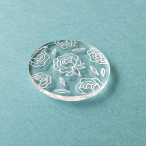 K&ouml;nnte beinhalten: Ein runder, transparenter Acryl-Anh&auml;nger mit einem wei&szlig;en Blumenmuster. Der Anh&auml;nger zeigt ein sich wiederholendes Muster aus Rosen und Bl&auml;ttern.