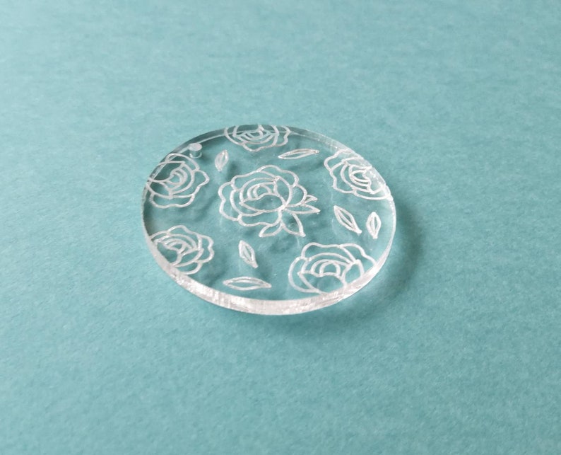 K&ouml;nnte beinhalten: Ein runder, transparenter Acryl-Anh&auml;nger mit einem wei&szlig;en Blumenmuster. Der Anh&auml;nger zeigt ein sich wiederholendes Muster aus Rosen und Bl&auml;ttern.