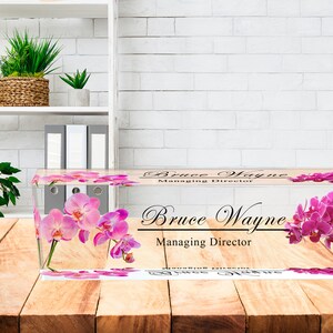 May include: A clear acrylic desk nameplate with a pink orchid design. The nameplate reads "Bruce Wayne, Managing Director".
