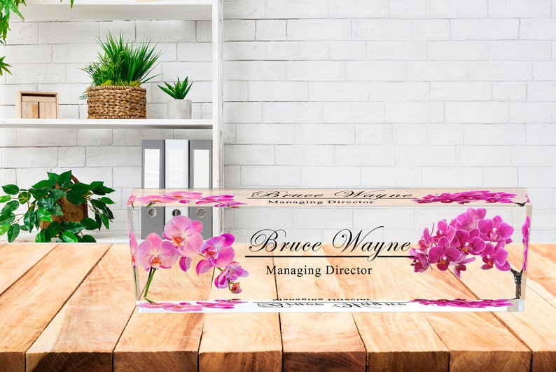 May include: A clear acrylic desk nameplate with a pink orchid design. The nameplate reads "Bruce Wayne, Managing Director".
