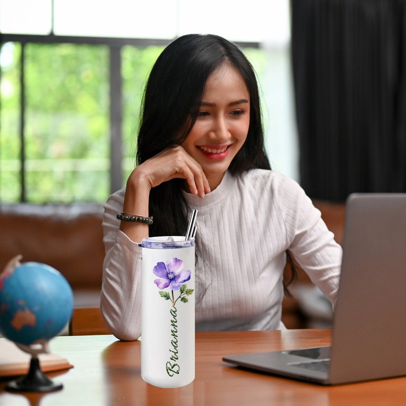 Puede incluir: Un vaso de acero inoxidable blanco con un dise&ntilde;o de flor morada y tallo verde. El vaso tiene una tapa transparente y una pajita. El nombre "Brianna" est&aacute; escrito verticalmente en el lateral del vaso.