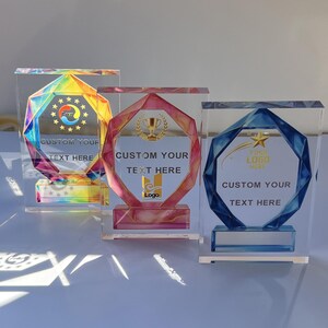 May include: Three clear acrylic trophies with geometric designs. One has a rainbow-colored design, another a pink and gold design, and the third a blue and gold design. Each trophy has space for custom text and a logo.