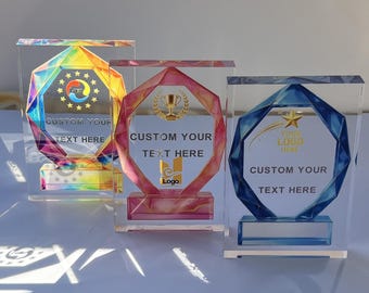 May include: Three clear acrylic trophies with geometric designs. One has a rainbow-colored design, another a pink and gold design, and the third a blue and gold design. Each trophy has space for custom text and a logo.