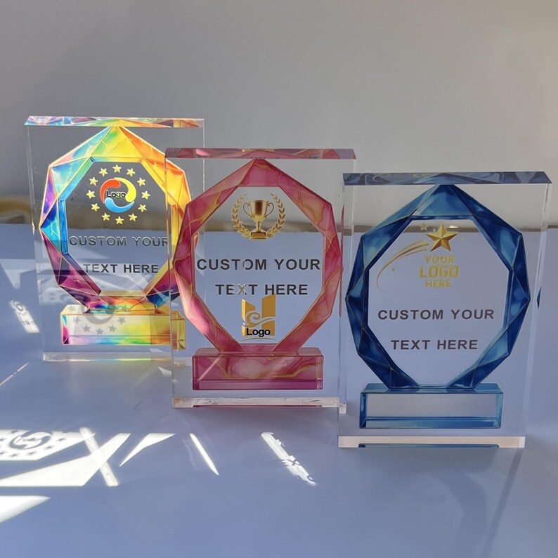 May include: Three clear acrylic trophies with geometric designs. One has a rainbow-colored design, another a pink and gold design, and the third a blue and gold design. Each trophy has space for custom text and a logo.