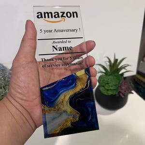 Puede incluir: Una placa de acr&iacute;lico transparente con un dise&ntilde;o abstracto azul y dorado. La placa tiene el texto "amazon" en negro, "5 year Amaversary!" en negro, "Awarded to Name" en negro y "Thank you for 5 years of service to Amazon!" en negro.