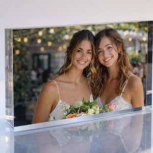 May include: A clear acrylic photo block displaying a color photograph of two smiling women. They are wearing white dresses and holding a bouquet of flowers. The background is blurred with soft lighting.
