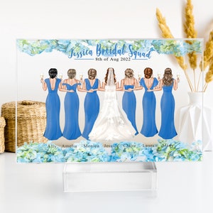 May include: A clear acrylic block with a blue floral design and a white background. The text "Jessica Bridal Squad" and "8th of Aug 2022" is printed on the block. There are seven bridesmaids in blue dresses and a bride in a white dress with a veil. The bridesmaids are holding champagne glasses. The names of the bridesmaids are printed below the image: Ally, Amelia, Monica, Jessica, Claire, Lauren, Macy.