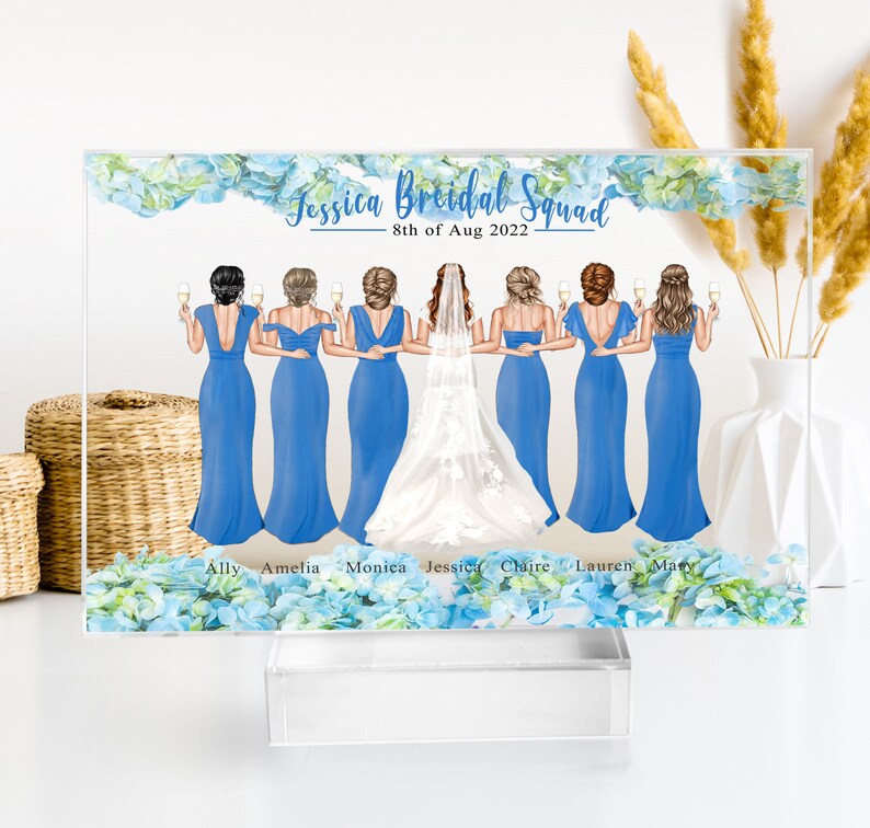 May include: A clear acrylic block with a blue floral design and a white background. The text "Jessica Bridal Squad" and "8th of Aug 2022" is printed on the block. There are seven bridesmaids in blue dresses and a bride in a white dress with a veil. The bridesmaids are holding champagne glasses. The names of the bridesmaids are printed below the image: Ally, Amelia, Monica, Jessica, Claire, Lauren, Macy.