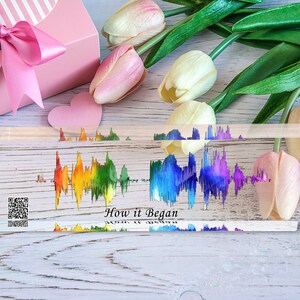 May include: A clear acrylic block with a rainbow-colored sound wave graphic and the text "How it Began". The block is on a white wooden surface with pink and white tulips and a pink gift box with a pink ribbon.
