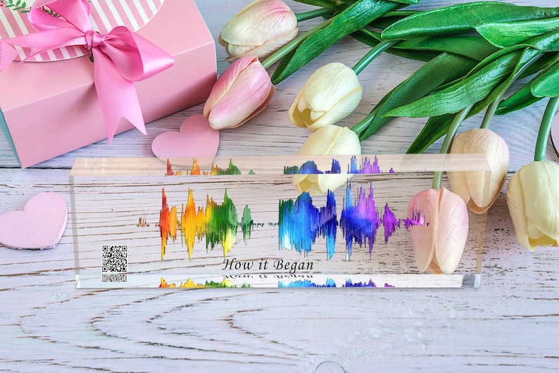 May include: A clear acrylic block with a rainbow-colored sound wave graphic and the text "How it Began". The block is on a white wooden surface with pink and white tulips and a pink gift box with a pink ribbon.