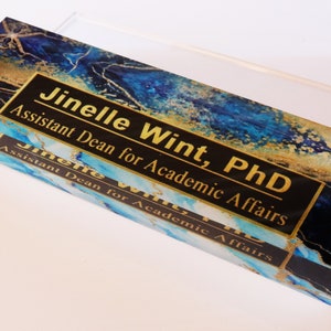 May include: A blue and gold acrylic desk nameplate with the text "Jinelle Wint, PhD Assistant Dean for Academic Affairs" in gold lettering on a black background.