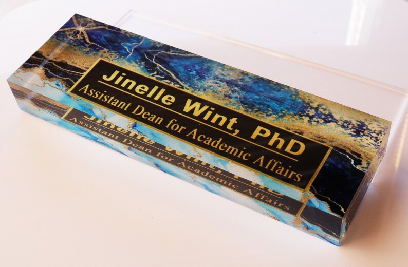 May include: A blue and gold acrylic desk nameplate with the text "Jinelle Wint, PhD Assistant Dean for Academic Affairs" in gold lettering on a black background.