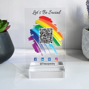 Könnte beinhalten: Ein klares Acrylschild mit dem Text "Let's Be Social" und einem regenbogenfarbenen Hintergrund. Ein QR-Code befindet sich in der Mitte des Schildes. Unter dem QR-Code befinden sich Social-Media-Icons für Facebook, LinkedIn, Twitter und Instagram. Der Text "@Tinacojewelry" befindet sich unter den Icons.