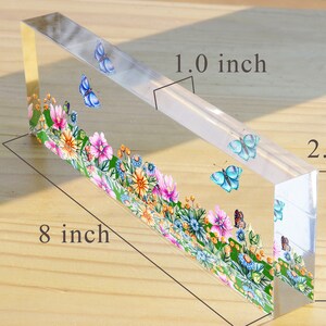 May include: A clear acrylic paperweight with a floral design and butterflies. The paperweight is 8 inches long, 2.5 inches wide, and 1 inch thick.