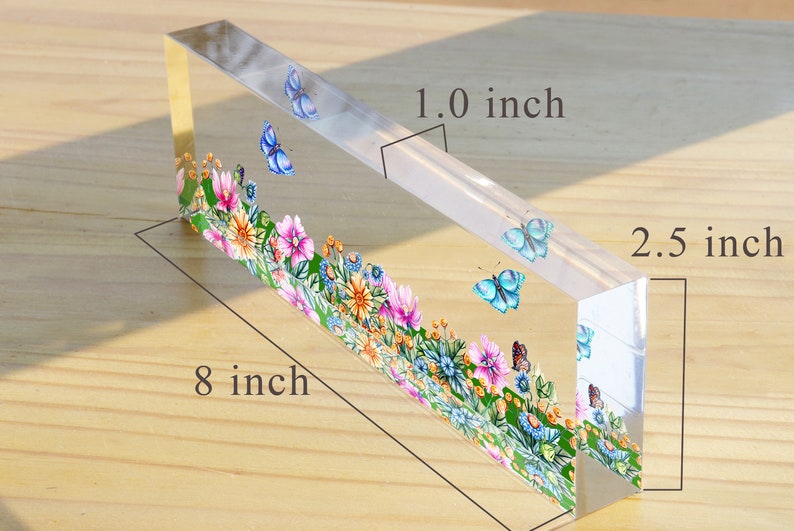 May include: A clear acrylic paperweight with a floral design and butterflies. The paperweight is 8 inches long, 2.5 inches wide, and 1 inch thick.