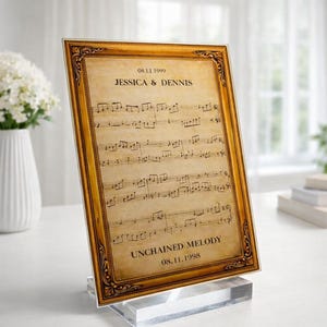Op de afbeelding: Een ingelijste print met bladmuziek met de tekst "JESSICA & DENNIS" en "UNCHAINED MELODY". De lijst heeft een goudkleurig, sierlijk ontwerp. De print wordt weergegeven op een heldere acryl standaard.