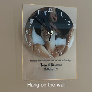 May include: A clear glass clock with a photo of a couple in bed. The clock face has black hands and black numbers. The text "Sharing time with you, love remains as first sight Tony & Brianna 8-09-2021" is printed below the clock face.