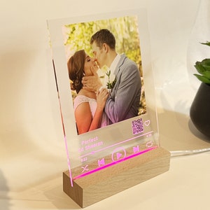 May include: A clear acrylic photo display with a wooden base. The display features a photo of a couple kissing, with the text "Perfect Ed Sheeran" and a music player interface design. The edges and music interface glow pink.