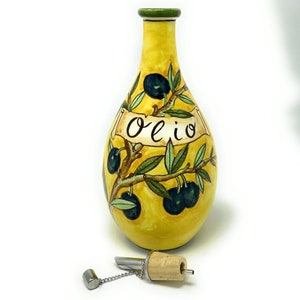 Może przedstawiać: Żółta ceramiczna butelka na oliwę z oliwek z zielonym korkiem i malowanym wzorem gałęzi oliwnej. Na butelce widnieje napis "Olio". Butelka jest wyposażona w korek z korka i metalowy łańcuch.