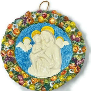 May include: A ceramic wall plaque depicting the Madonna and Child surrounded by a wreath of fruit and flowers. The plaque is painted in vibrant colors, including blue, yellow, green, and white.