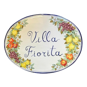 Puede incluir: Letrero ovalado de cerámica con las palabras "Villa Fiorita" en escritura azul. El letrero está decorado con un borde de frutas y flores coloridas, incluyendo limones, uvas y naranjas. El letrero tiene un borde azul.