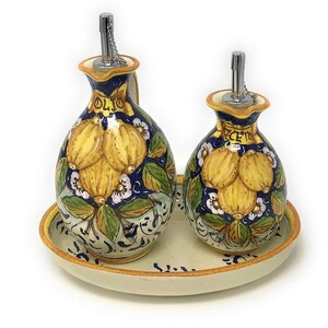 May include: A set of two ceramic oil and vinegar bottles with a lemon design. The bottles are decorated with yellow lemons, green leaves, white flowers, and blue accents. The bottles are on a white oval tray with a brown rim.