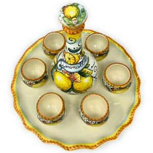 May include: A ceramic limoncello set featuring a bottle and six small cups, all decorated with yellow lemons and green leaves. The bottle has a cork stopper and the word "limoncello" on a banner. The set sits on a scalloped tray with a yellow and blue border.