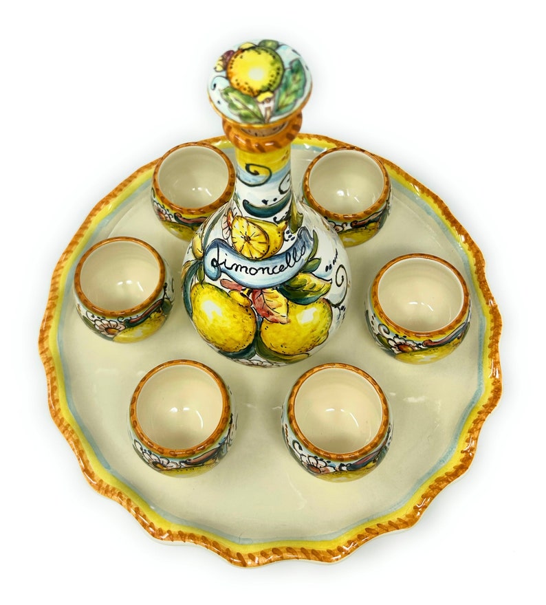 May include: A ceramic limoncello set featuring a bottle and six small cups, all decorated with yellow lemons and green leaves. The bottle has a cork stopper and the word "limoncello" on a banner. The set sits on a scalloped tray with a yellow and blue border.