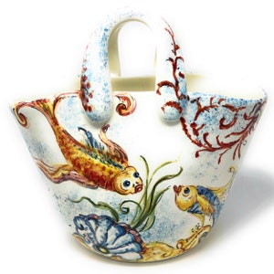 May include: A white ceramic hand-painted vase shaped like a handbag with a handle. The vase features a colorful design of fish, flowers, and foliage.