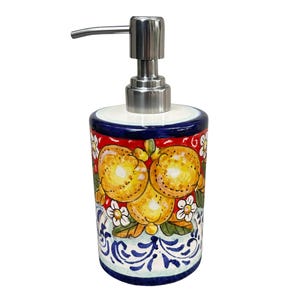 Op de afbeelding: Een keramische zeepdispenser met een roestvrijstalen pomp. De cilindrische behuizing is versierd met een levendig ontwerp met gele vruchten, witte bloemen en blauw-witte krullen tegen een rode achtergrond. De rand is blauw.