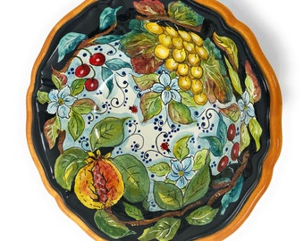 May include: A round ceramic bowl with a black background and a colorful floral design. The bowl features a variety of fruits and flowers, including grapes, cherries, and pomegranates. The bowl has a scalloped edge and a gold rim.