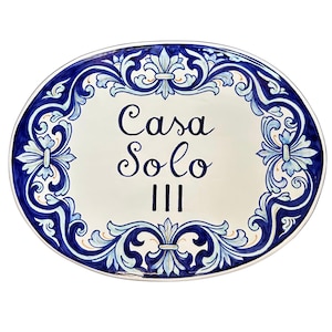 May include: An oval ceramic plaque featuring a white center with the dark blue text Casa Solo III. The border is decorated with intricate blue and light blue floral scrollwork patterns on a white background, creating a decorative sign.
