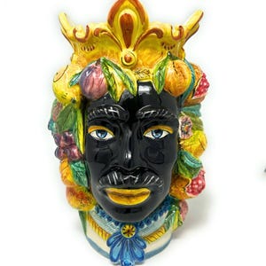 May include: A black ceramic vase shaped like a human head with a crown of fruit and flowers. The vase has a yellow crown, blue eyes, and a yellow mustache.