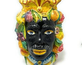 May include: A ceramic head-shaped vase featuring a glossy black face with blue eyes and a mustache. The head is adorned with a yellow crown, green leaves, and various colorful fruits including grapes, oranges, and berries, with a blue floral collar.