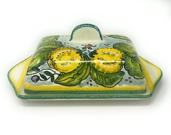 May include: A ceramic butter dish with a lid featuring a hand-painted design of lemons and leaves on a light blue background. The dish has a green and yellow border.