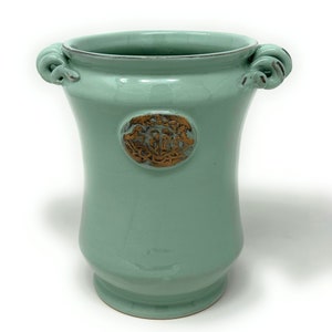 Puede incluir: Un jarrón de cerámica verde claro con dos asas y un escudo dorado en relieve. El jarrón tiene un fondo redondeado y una parte superior acampanada.